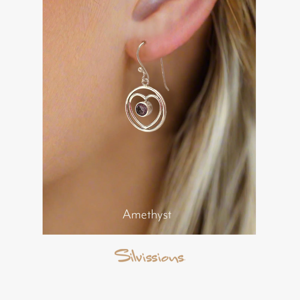 A pair of sterling silver heart-shaped dangle earrings with an amethyst gemstone, hanging from a person's ear.