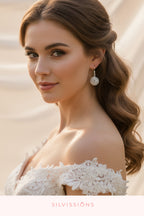 Woman wearing a white lace off-shoulder top with earrings, standing against a soft blurred background.