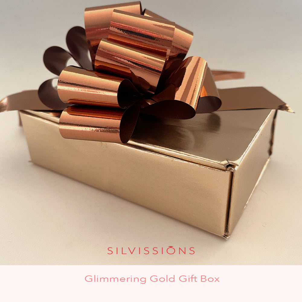A close-up of a gift box wrapped in metallic rose-gold foil with a matching large bow, on a light background. Text at the bottom reads 'SILVISSIONS' and 'Glimmering Gold'.