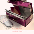 A purple and gold Christmas-themed gift box with a holly tree design, shown open with wrapped silver gift bags and a small card inside. A small plastic bag with a handwritten note sits beside it on a light surface.