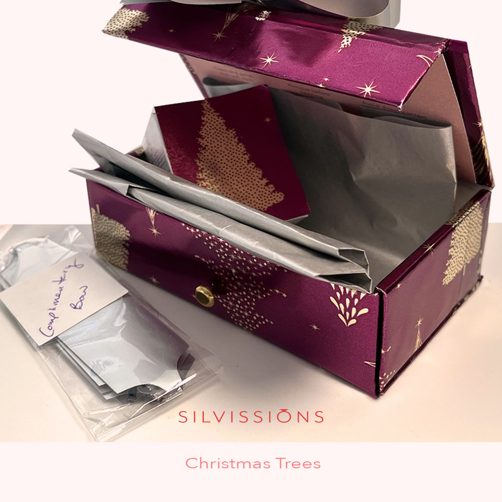 A purple and gold Christmas-themed gift box with a holly tree design, shown open with wrapped silver gift bags and a small card inside. A small plastic bag with a handwritten note sits beside it on a light surface.