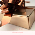 A close-up of a gift box wrapped in metallic rose-gold foil with a matching large bow, on a light background. Text at the bottom reads 'SILVISSIONS' and 'Glimmering Gold'.