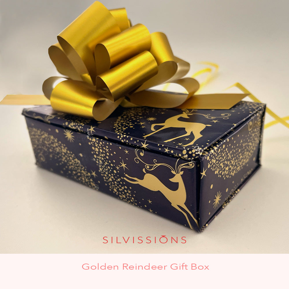 A square gift box wrapped in dark navy blue paper with a gold reindeer and star pattern, topped with a large shiny gold ribbon bow. The text 'SILVISSIÕNS' and 'Golden Reindeer Gift Box' appears below.