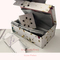 An open decorative gift box with a white exterior patterned with gold, black, and pink snowflake motifs, filled with silver envelopes and a small card. A clear plastic bag with a handwritten note lies beside it on a light surface.