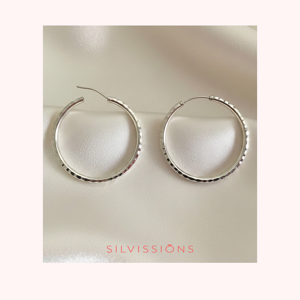 Pair of silver hoop earrings on a blue fabric background with 'Silvissions' branding.