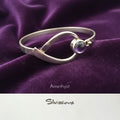 Silver bangle bracelet with a purple gemstone on a soft pink background with white roses.