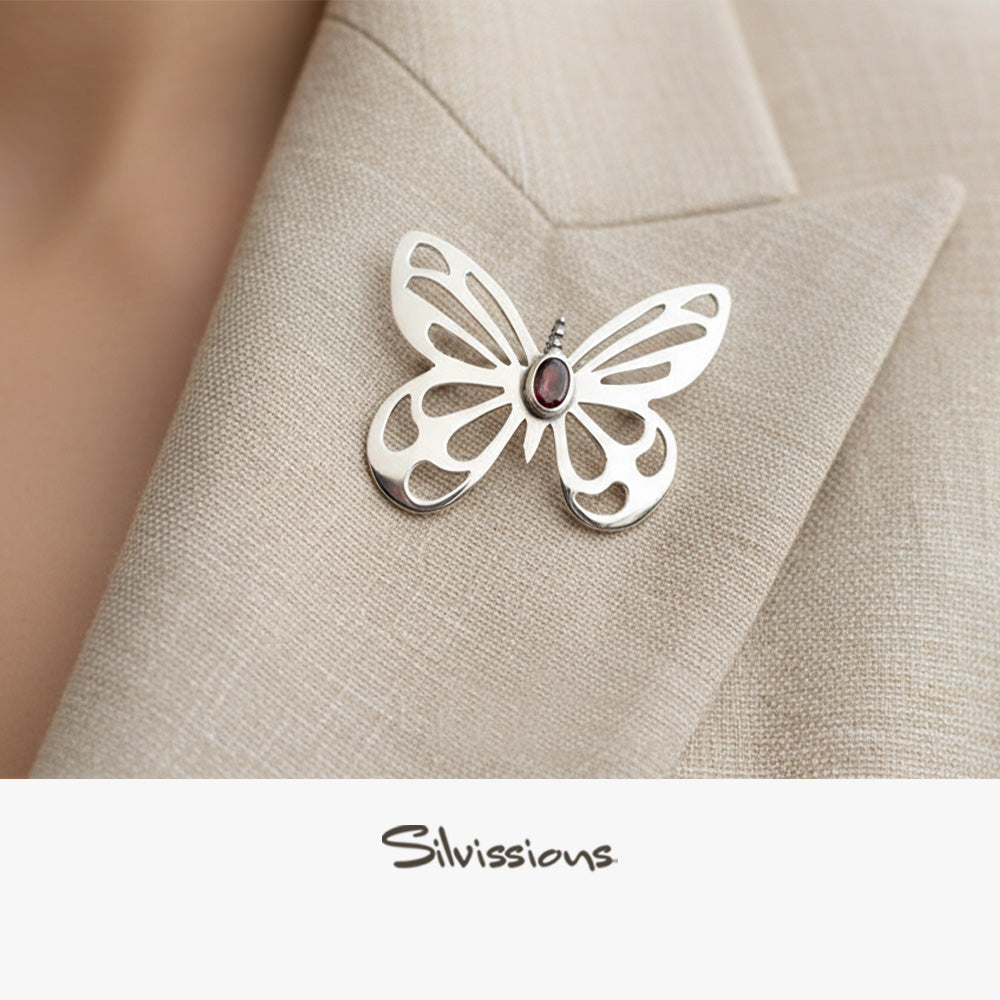 Silver butterfly brooch with a red gemstone on a beige fabric background, branded 'Silvissions'.