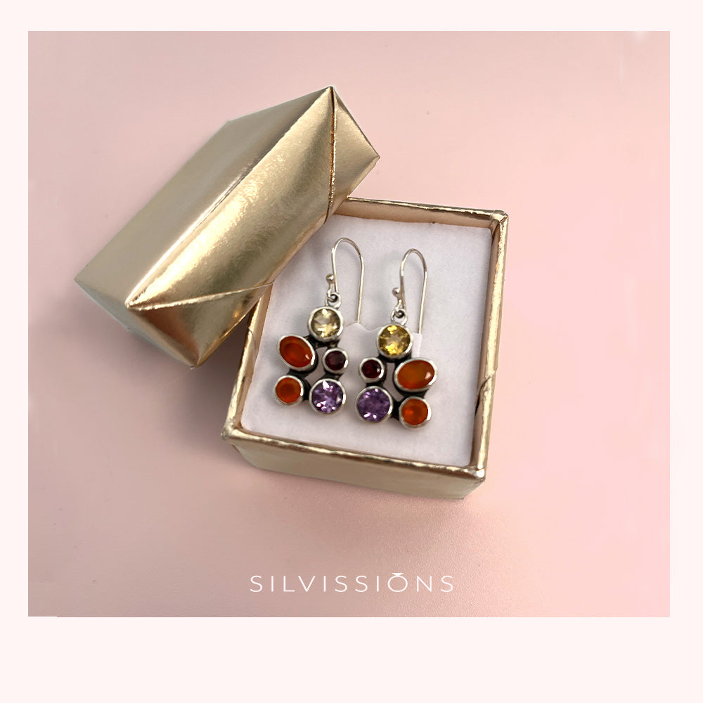 A pair of colorful gemstone cluster dangle earrings displayed in an open metallic gold jewelry box on a soft pink background. The earrings feature multiple round gemstones in orange, purple, yellow, and red tones set in silver-toned metal.