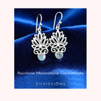 Silver earrings with rainbow moonstone stones on a blue satin background, featuring the brand 'Silvissions'.