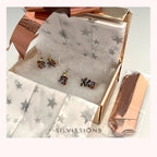 A gift box containing silver-toned earrings and a matching pendant with colorful gemstone accents, lined up on white star-patterned tissue paper. A metallic rose gold box is partially open, and a small packaged item sits beside it.