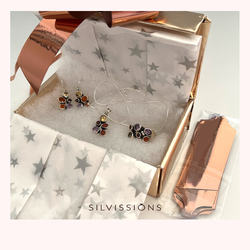 A gift box containing silver-toned earrings and a matching pendant with colorful gemstone accents, lined up on white star-patterned tissue paper. A metallic rose gold box is partially open, and a small packaged item sits beside it.