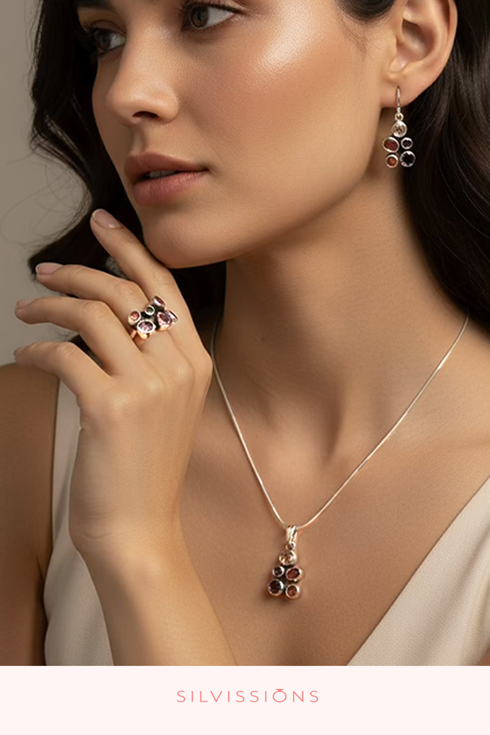 A close-up lifestyle shot of a model wearing a silver-toned jewelry set from the brand SILVISSIONS, featuring a delicate pendant necklace, matching drop earrings, and a ring with dark pinkish-red stones.