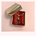 A silver pendant necklace with multiple colorful gemstones is displayed inside an open gold jewelry box lined with red satin. The brand name 'SILVISSIONS' is printed in red at the bottom of the image against a light pink background.
