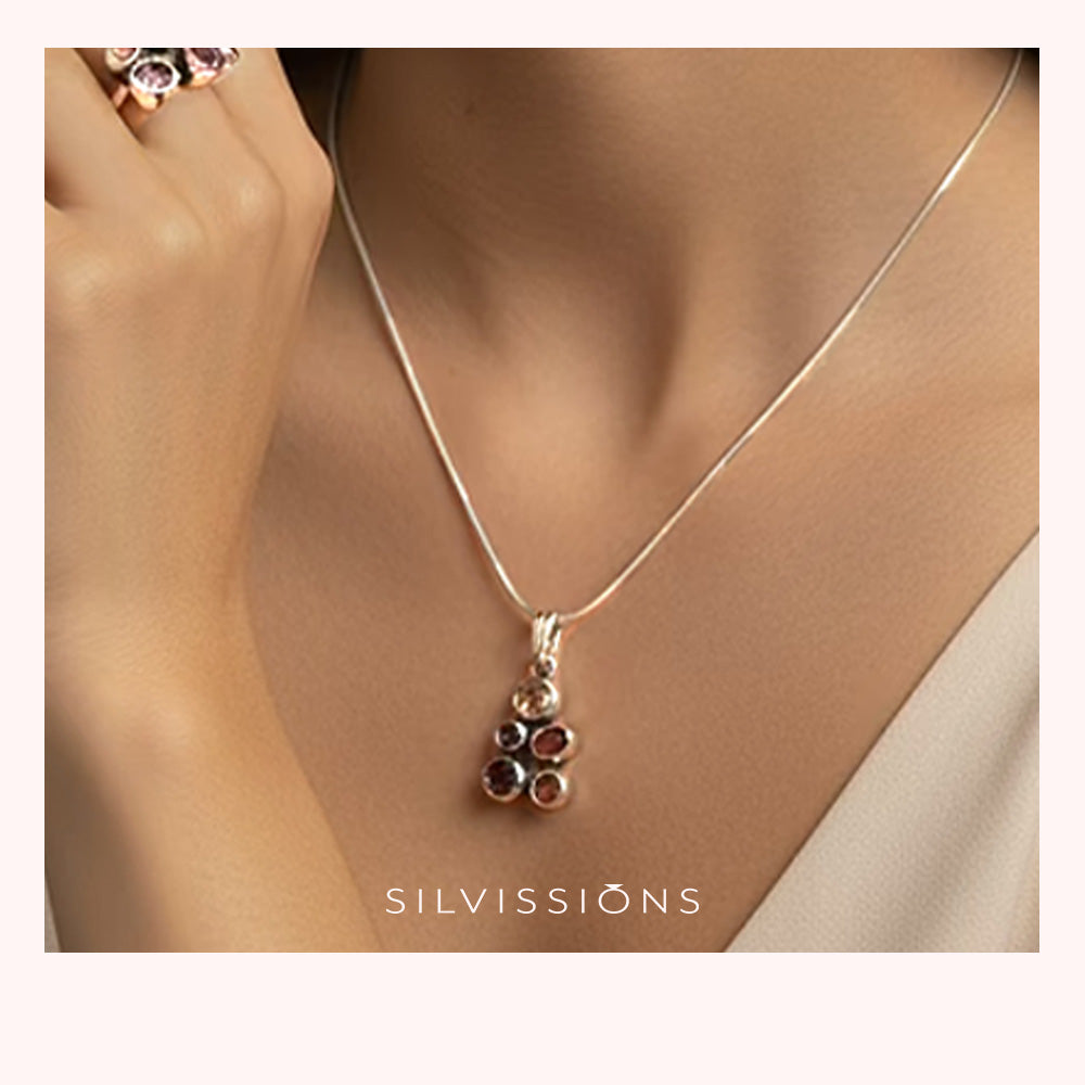A close-up lifestyle shot of a model wearing a delicate silver chain necklace with a small pendant featuring dark red gemstones. The model's neck and chest are visible, wearing a light-colored top, with a ring on her finger.