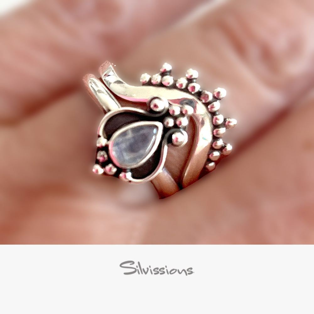 Silver ring with a central rainbow moonstone gemstone held between fingers, brand 'Silvissions' visible.