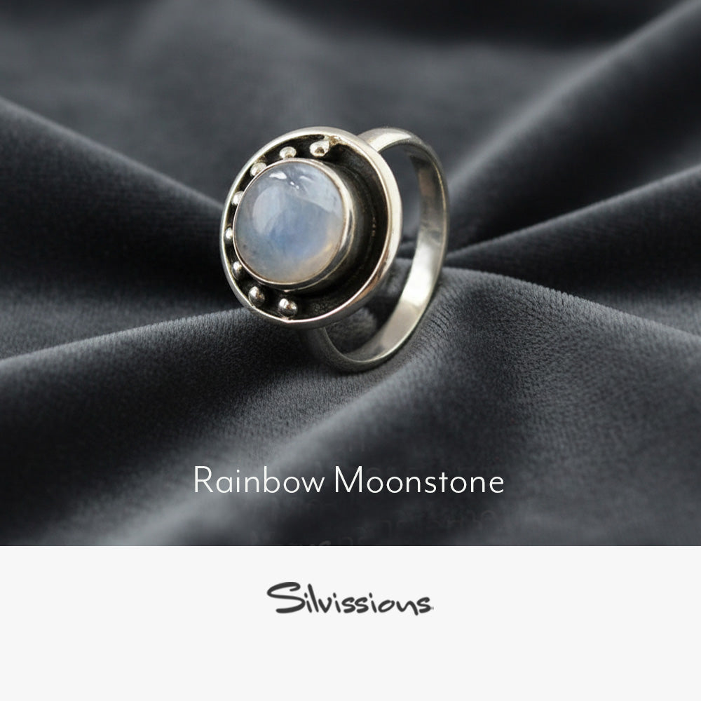 Silver ring with a rainbow moonstone on a dark fabric background, featuring the brand 'Silvissions'.