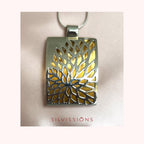 A silver-toned rectangular pendant necklace with a tree-of-life cutout design showing a gold-toned interior, suspended from a thin silver chain against a pale pink background. The brand name 'SILVISSIONS' is printed in red text below the pendant.