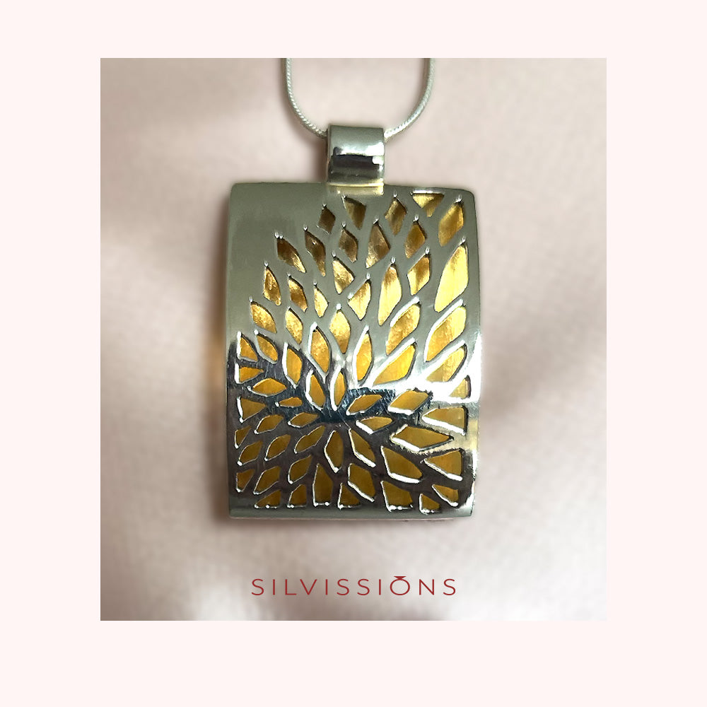 A silver-toned rectangular pendant necklace with a tree-of-life cutout design showing a gold-toned interior, suspended from a thin silver chain against a pale pink background. The brand name 'SILVISSIONS' is printed in red text below the pendant.