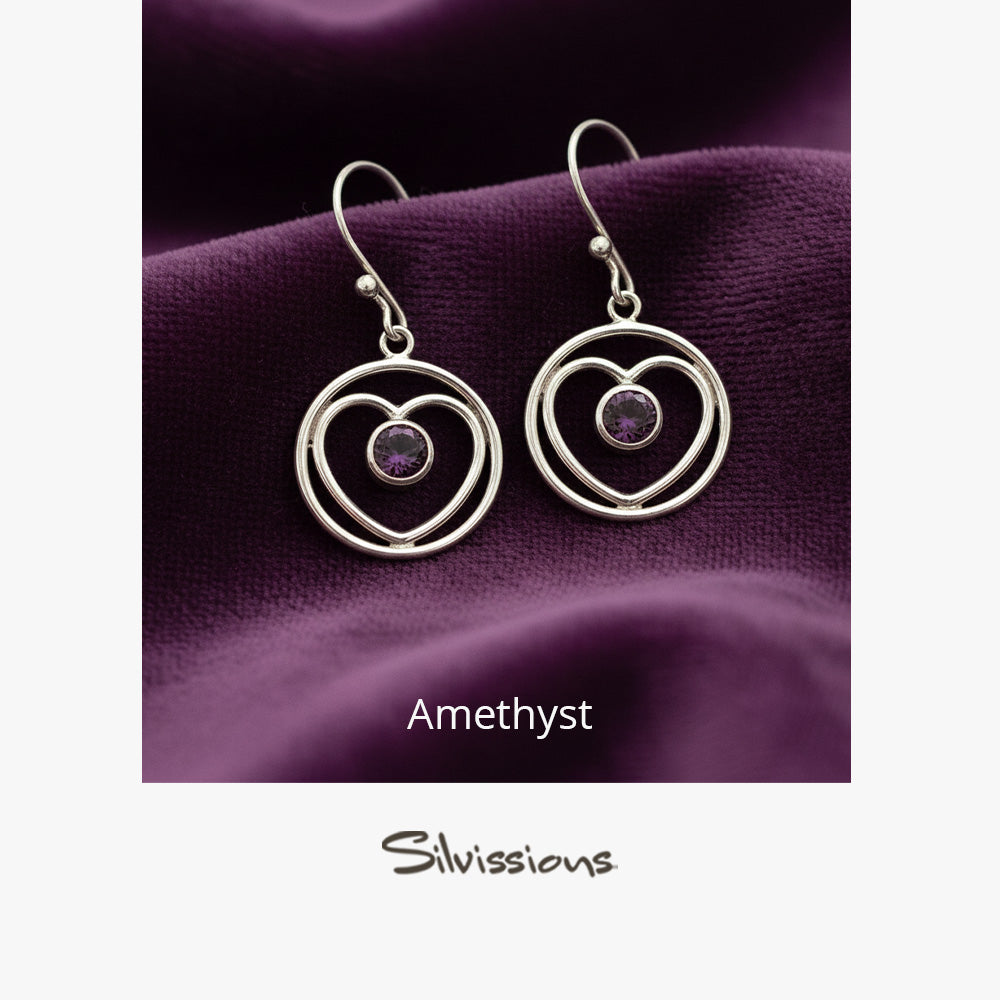 Silver heart-shaped earrings with purple gemstones on a purple fabric background, featuring the brand 'Silvissions'.
