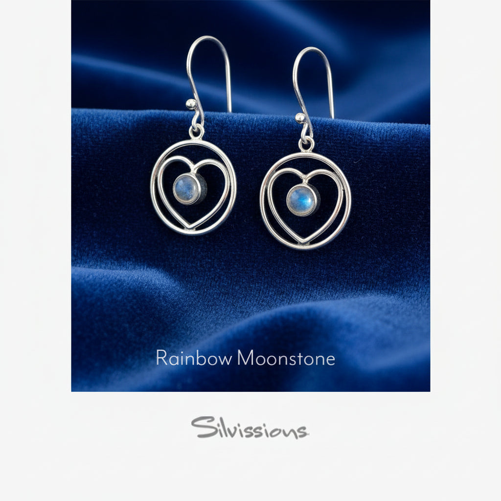 Heart-shaped earrings with rainbow moonstone stones on a blue satin background, branded 'Silvissions'.