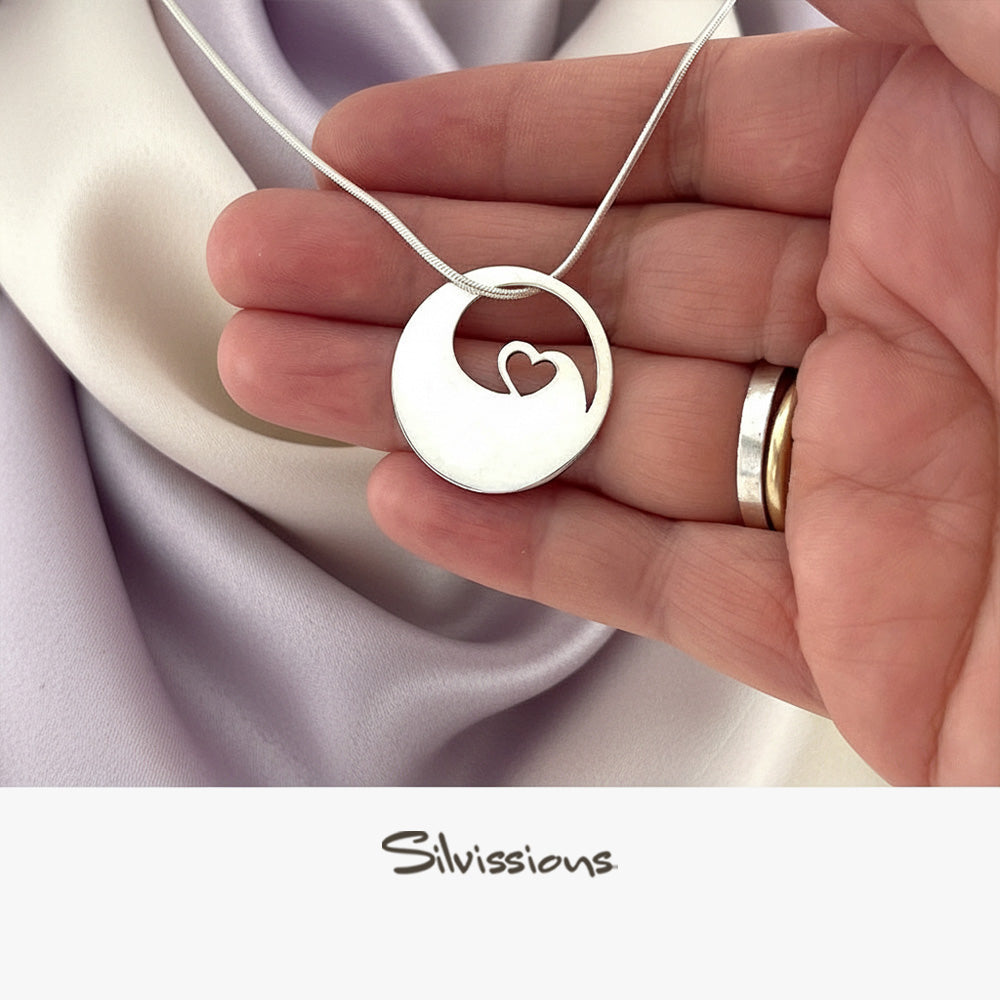 Silver necklace with a heart pendant held by a hand on a soft fabric background, featuring the brand 'Silvissions'.