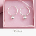 Silver hoop earrings with heart charms on a pink fabric background, featuring the brand 'Silvissions'.