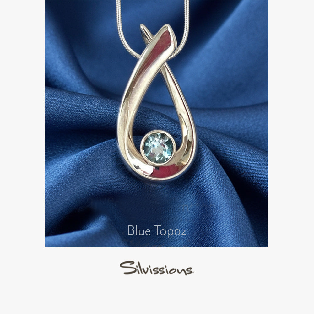 Silver pendant with blue gemstone on blue fabric, featuring 'Silvissions' brand.