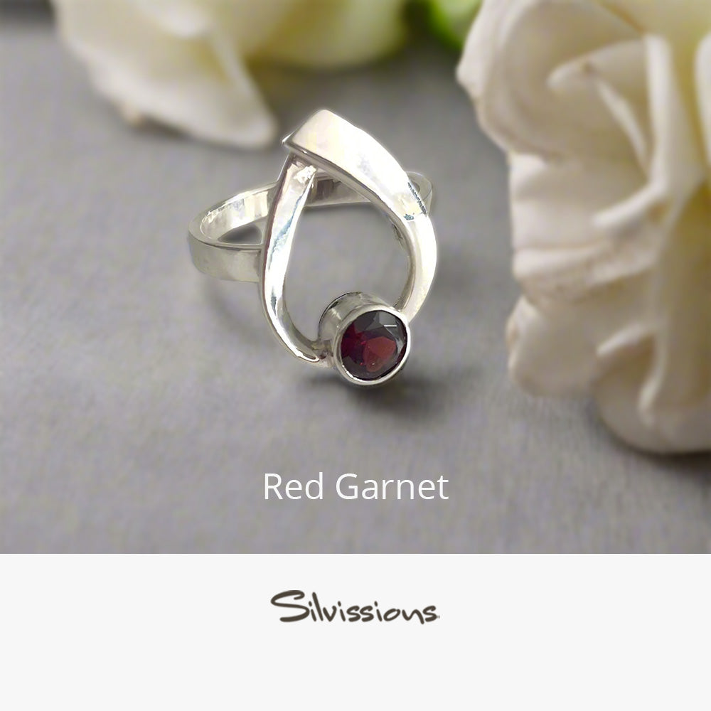 Silver ring with a red gemstone on a gray surface with flowers in the background, featuring the brand 'Silvissions'.