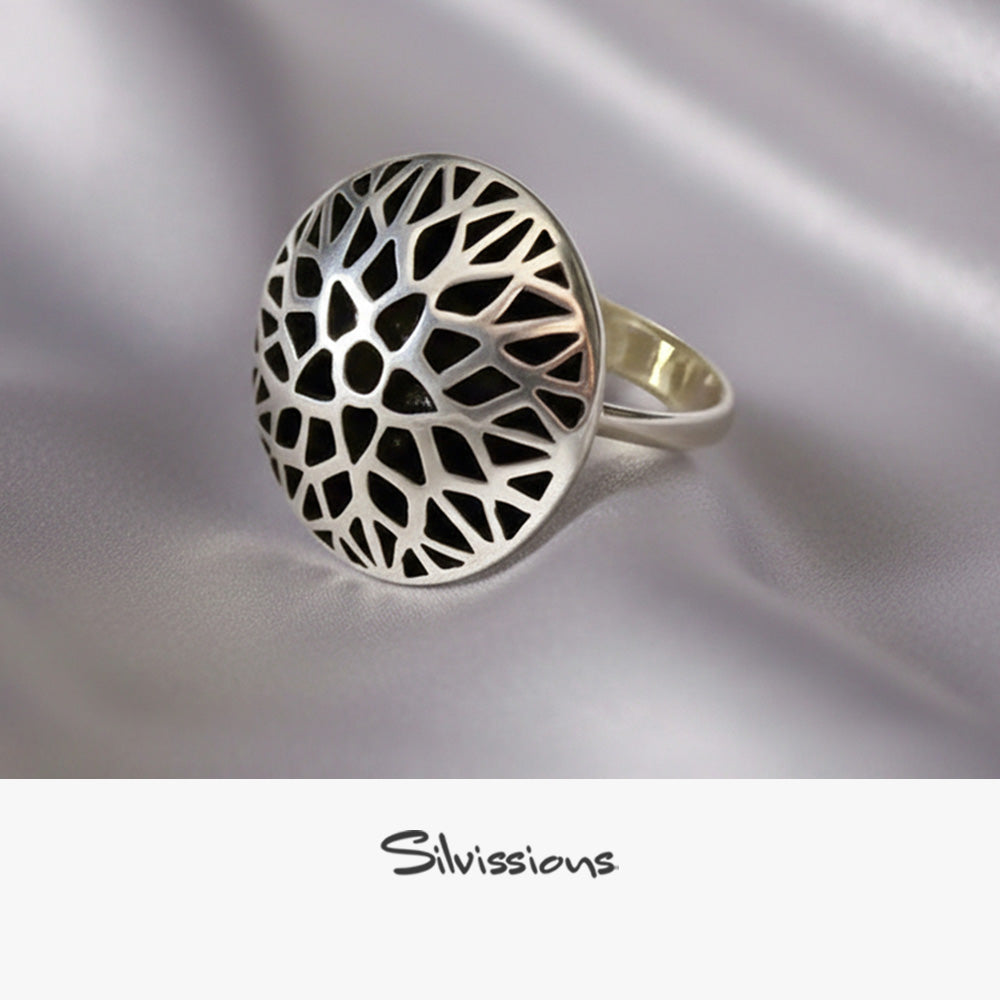 Silver ring with intricate design on a gray surface with white flowers in the background, featuring the brand 'Silvissions'.