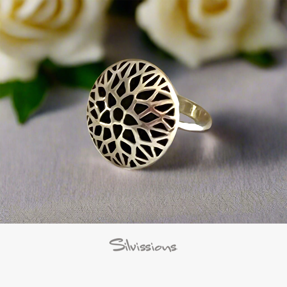 Silver ring with intricate design on a gray surface with white flowers in the background, featuring the brand 'Silvissions'.
