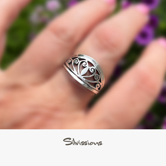 Silver ring with intricate design on a hand, blurred floral background, Silvissions branding.