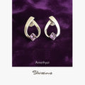 Pair of silver earrings with amethyst gemstones on a purple fabric background, branded 'Silvissions'.