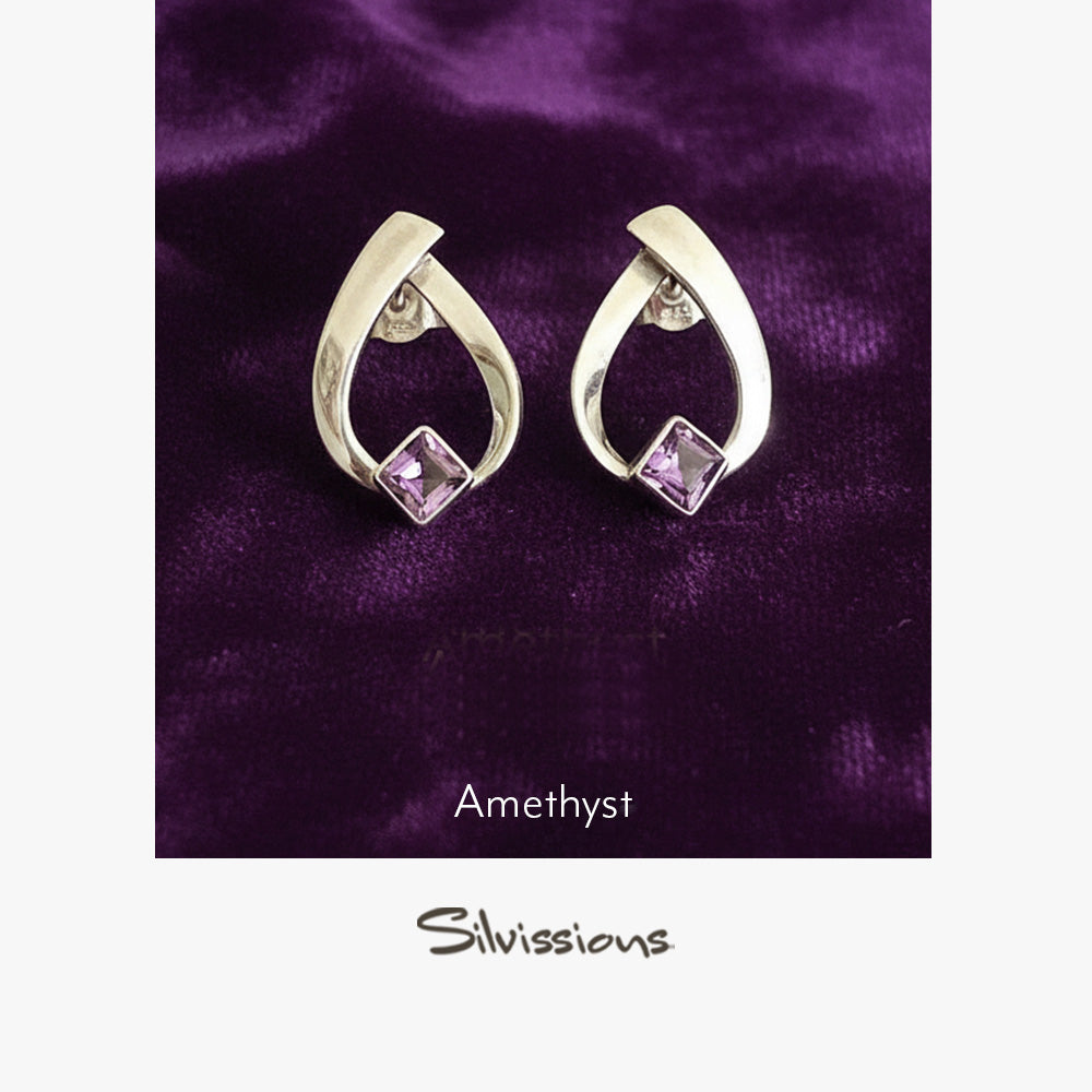 Pair of silver earrings with amethyst gemstones on a purple fabric background, branded 'Silvissions'.