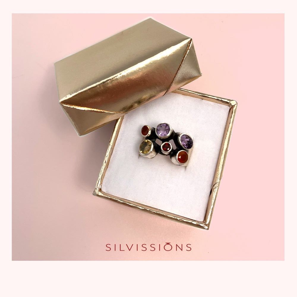 A cluster of small gemstone stud earrings in silver settings, displayed inside an open square jewelry box with a metallic gold lid, on a soft pink background. The brand name 'SILVISSIONS' is printed at the bottom.