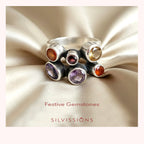 A silver ring with six bezel-set gemstones in amber, purple, and yellow tones, displayed on draped champagne satin fabric. The brand name 'SILVISSIONS' and the text 'Festive Gemstones' are printed below the ring.